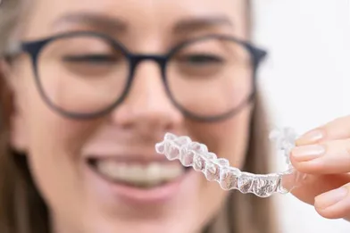 A woman smiling while holding a clear aligner for her teeth.